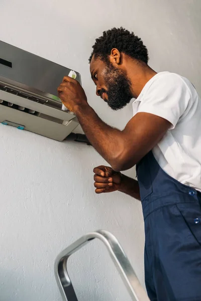 Atento reparador afroamericano de pie en la escalera y la fijación de aire acondicionado - foto de stock