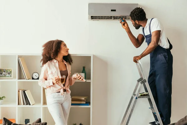 Africano americano reparador fixando ar condicionado perto de mulher americana africana bonita segurando controlador remoto — Fotografia de Stock