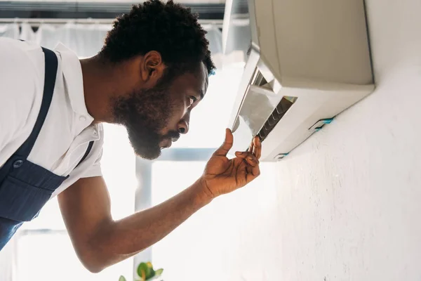 Concentrated african american repairman looking at broken air conditioner — Stock Photo