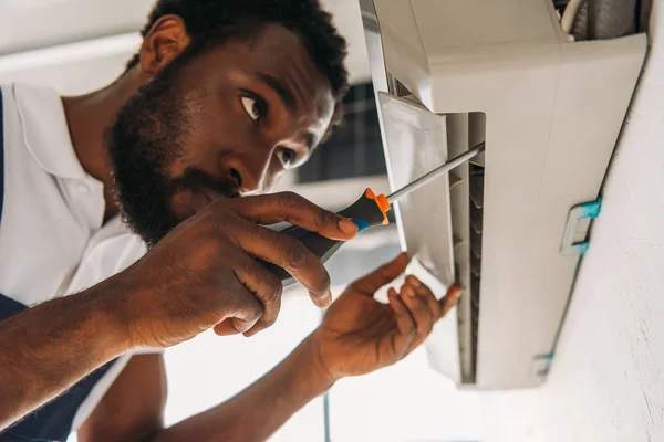 Réparateur afro-américain sérieux fixant climatiseur avec tournevis — Photo de stock