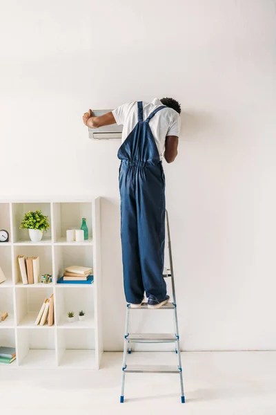 Rückansicht eines afrikanisch-amerikanischen Reparateurs, der auf einer Leiter steht und eine Klimaanlage befestigt — Stockfoto