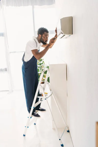 Vista posterior del reparador afroamericano de pie en la escalera y la fijación de aire acondicionado - foto de stock