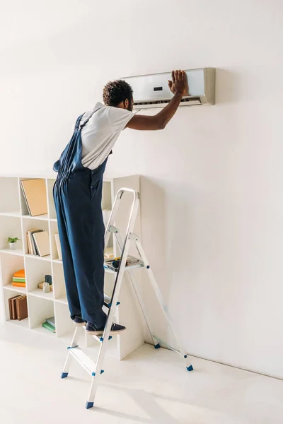 Африканский рабочий в комбинезоне, стоящий на лестнице и ремонтирующий кондиционер — стоковое фото