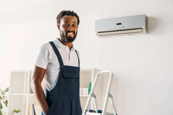 Joyeux réparateur afro-américain debout sur l'échelle près du climatiseur et souriant à la caméra — Photo de stock