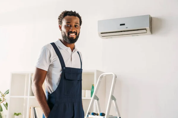 Alegre Africano americano reparador de pé na escada perto de ar condicionado e sorrindo para a câmera — Fotografia de Stock