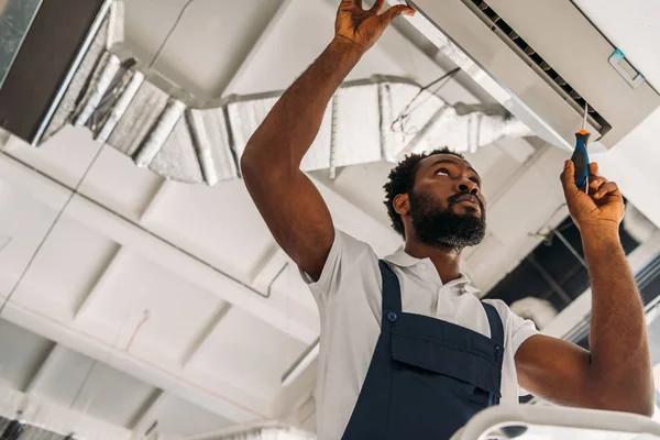 Visão de baixo ângulo de afro americano handyman reparando ar ar condicionado com chave de fenda — Fotografia de Stock