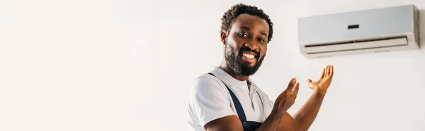 Panoramaaufnahme eines glücklichen afrikanisch-amerikanischen Reparateurs, der in die Kamera lächelt und mit den Händen auf die Klimaanlage zeigt — Stockfoto