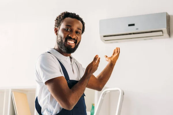 Cheerful african american repairman smiling at camera and pointing with hands at air conditioner — Stock Photo