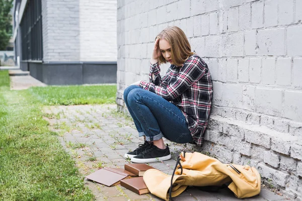 Triste et blonde adolescent en chemise et jeans assis près du mur parmi les livres — Photo de stock