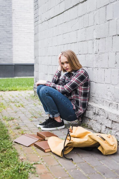 Déçu et blond adolescent en chemise et jeans assis près du mur parmi les livres — Photo de stock