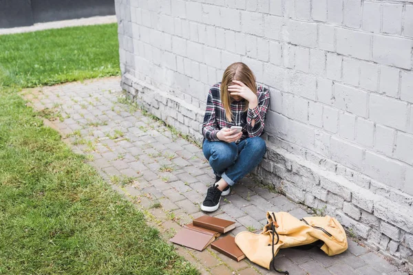 Разочарованный и блондинка подросток в рубашке и джинсах сидит у стены и держит смартфон — стоковое фото
