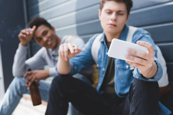 Selective focus of friends smiling, smoking cigarettes, holding beer and taking selfie — Stock Photo