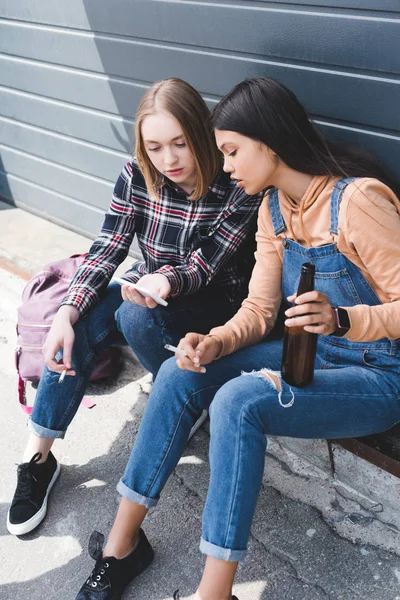 Bonitos amigos fumando cigarrillos, sosteniendo cerveza, sentado y mirando el teléfono inteligente - foto de stock