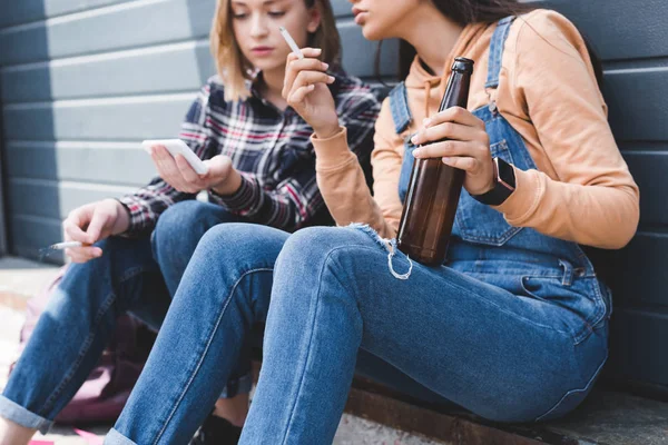 Enfoque selectivo de amigos bonitos fumando cigarrillos, sosteniendo cerveza, sentado y mirando el teléfono inteligente - foto de stock