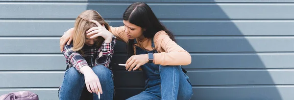 Tiro panorâmico de adolescentes tristes e bonitos sentado e segurando cigarros — Fotografia de Stock