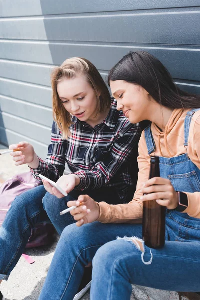 Pretty friends smoking cigarettes, holding beer, sitting and looking at smartphone — Stock Photo
