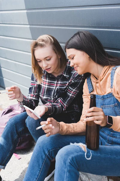 De jolis amis fumant des cigarettes, tenant de la bière, assis et regardant smartphone — Photo de stock