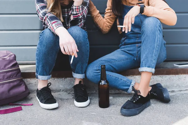 Cropped view of teens smoking cigarettes, holding beer and sitting — Stock Photo