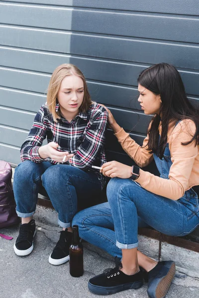 Adolescentes decepcionados y bonitos sentados, hablando y sosteniendo cigarrillos - foto de stock