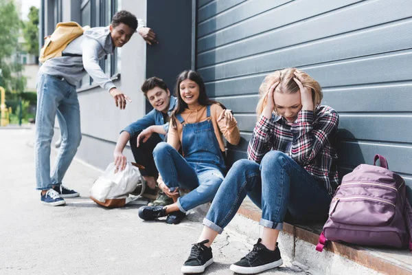 Amigos sonrientes acosando y señalando con los dedos a la triste rubia adolescente - foto de stock