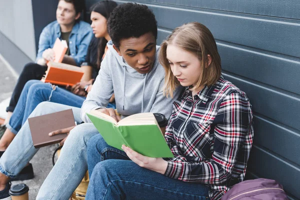 African american and caucasian teenagers sitting, talking and reading book — Stock Photo