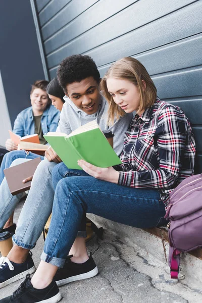 African american and caucasian teenagers sitting, talking and reading book — Stock Photo