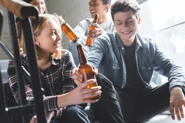 Adolescentes sonrientes sentados en las escaleras, bebiendo cerveza y hablando - foto de stock