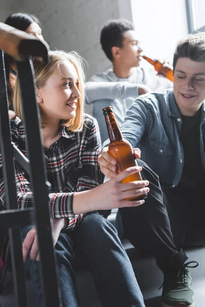 Adolescentes sonrientes sentados en las escaleras, sosteniendo la cerveza y hablando - foto de stock
