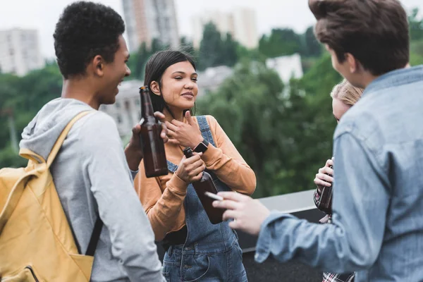 Adolescentes sonrientes sosteniendo botellas de vidrio con cerveza y fumar cigarrillos - foto de stock