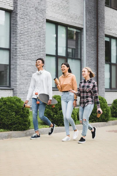 Adolescentes sonrientes caminando, sosteniendo vasos desechables y mirando hacia otro lado - foto de stock