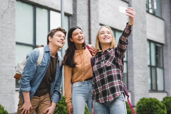 Adolescentes felices sosteniendo el teléfono inteligente, tomando selfie y sonriendo fuera - foto de stock