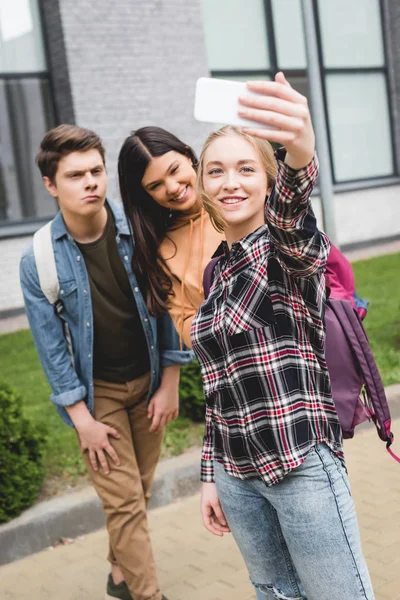 Positive teenagers holding smartphone, taking selfie and smiling outside — Stock Photo