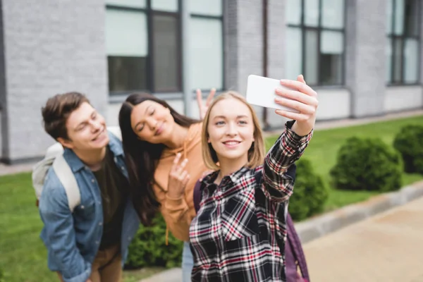 Щасливі підлітки тримають смартфон, беручи селфі і посміхаючись надворі — Stock Photo