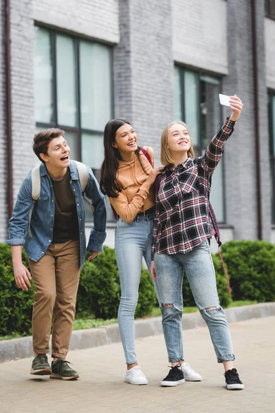 Adolescentes felices sosteniendo el teléfono inteligente, tomando selfie y sonriendo fuera - foto de stock