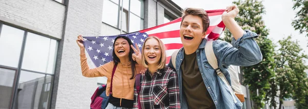 Foto panorámica de adolescentes felices sonriendo, sosteniendo la bandera americana y mirando a la cámara - foto de stock