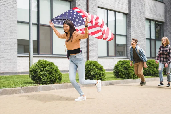 Adolescentes felices sonriendo, sosteniendo la bandera americana y corriendo afuera - foto de stock