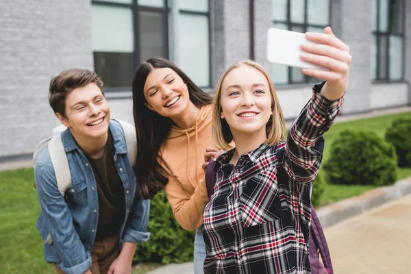 Adolescentes alegres sosteniendo el teléfono inteligente, tomando selfie y sonriendo fuera - foto de stock