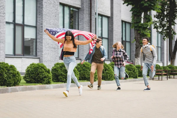Adolescentes felices sonriendo, sosteniendo la bandera americana y corriendo afuera — Stock Photo