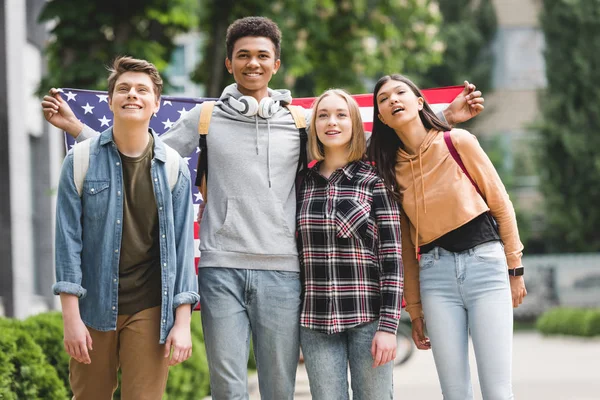 Adolescentes felices sosteniendo bandera americana, sonriendo y mirando hacia otro lado - foto de stock