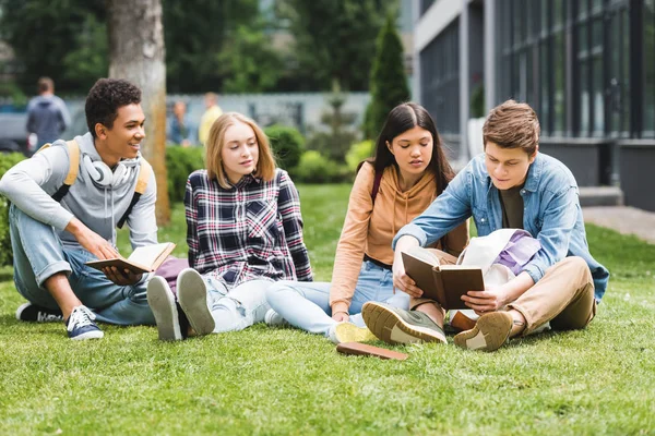 Lächelnde Teenager, die auf Gras sitzen und draußen Bücher lesen — Stockfoto