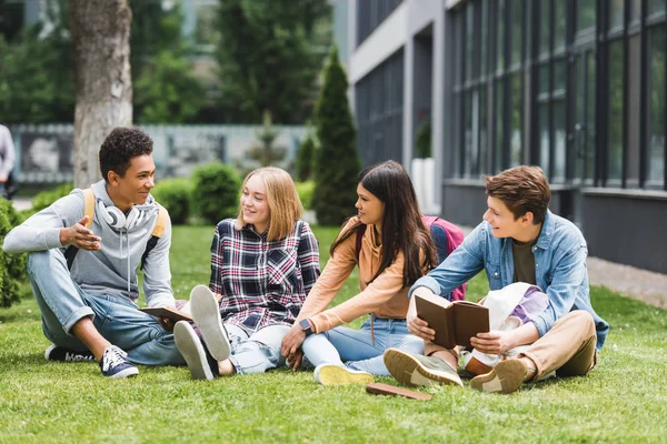 Adolescenti sorridenti seduti sull'erba, che parlano e tengono libri — Foto stock