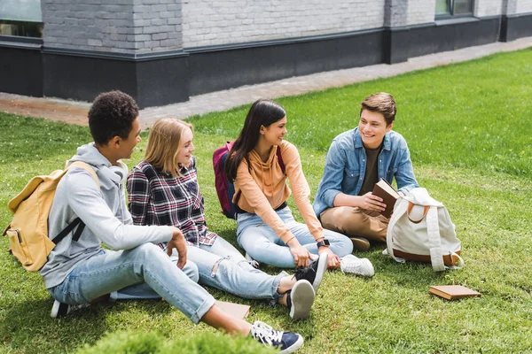 Smiling teenagers sitting on grass, talking, holding book outside — Stock Photo
