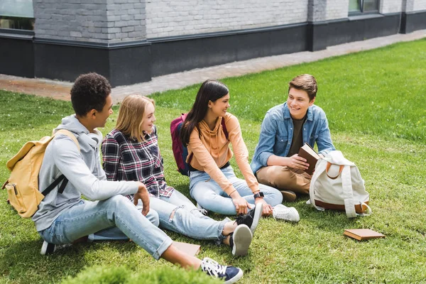 Adolescentes sonrientes sentados en la hierba, hablando, sosteniendo el libro afuera - foto de stock