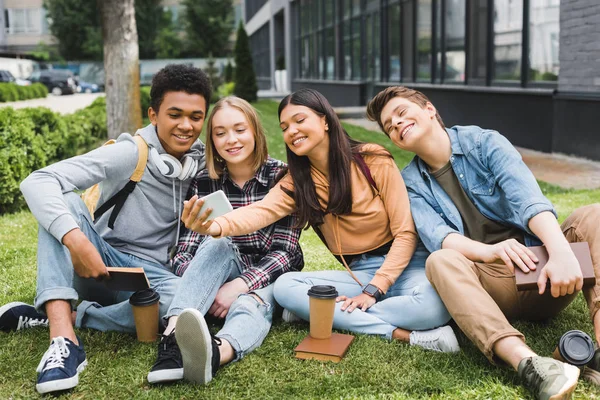 Lächelnde und glückliche Teenager, die auf Gras sitzen und ein Selfie machen — Stockfoto