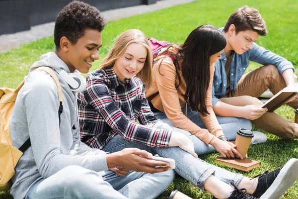 Sorrindo e felizes adolescentes sentados na grama, olhando para smartphone e livro de leitura — Fotografia de Stock
