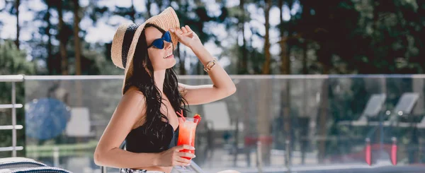 Красива брюнетка молода жінка в купальнику, солом'яний капелюх і сонцезахисні окуляри посміхається, тримаючи коктейль, панорамний знімок — стокове фото