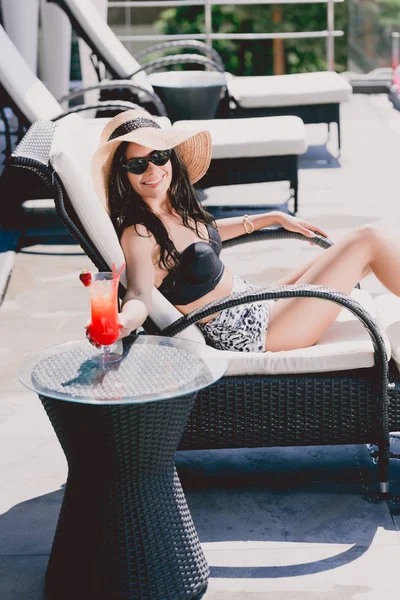 Glücklich schöne brünette junge Frau in Badeanzug, Strohhut und Sonnenbrille sonnt sich und hält eisigen Erdbeercocktail auf dem Tisch — Stockfoto