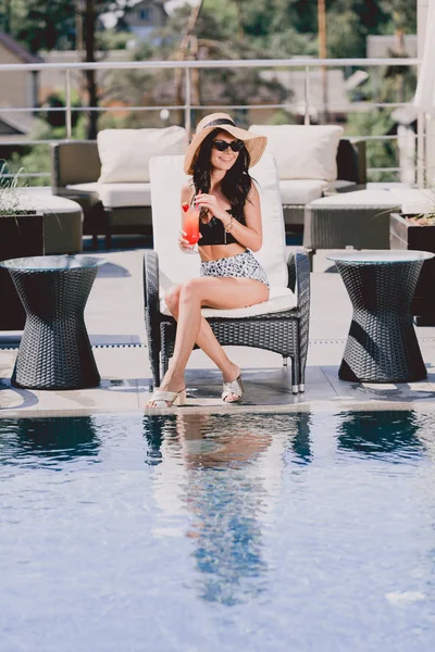 Beautiful brunette young woman in swimsuit, straw hat and sunglasses smiling and looking away while holding cocktail near swimming pool — Stock Photo