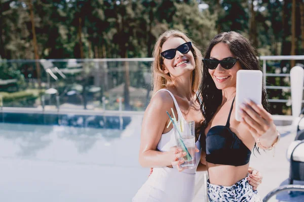 Morena sexy y sonriente y mujeres rubias en trajes de baño tomando selfie cerca de la piscina - foto de stock