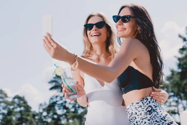 Vue à angle bas des femmes brunes et blondes heureuses en maillots de bain prenant selfie — Photo de stock